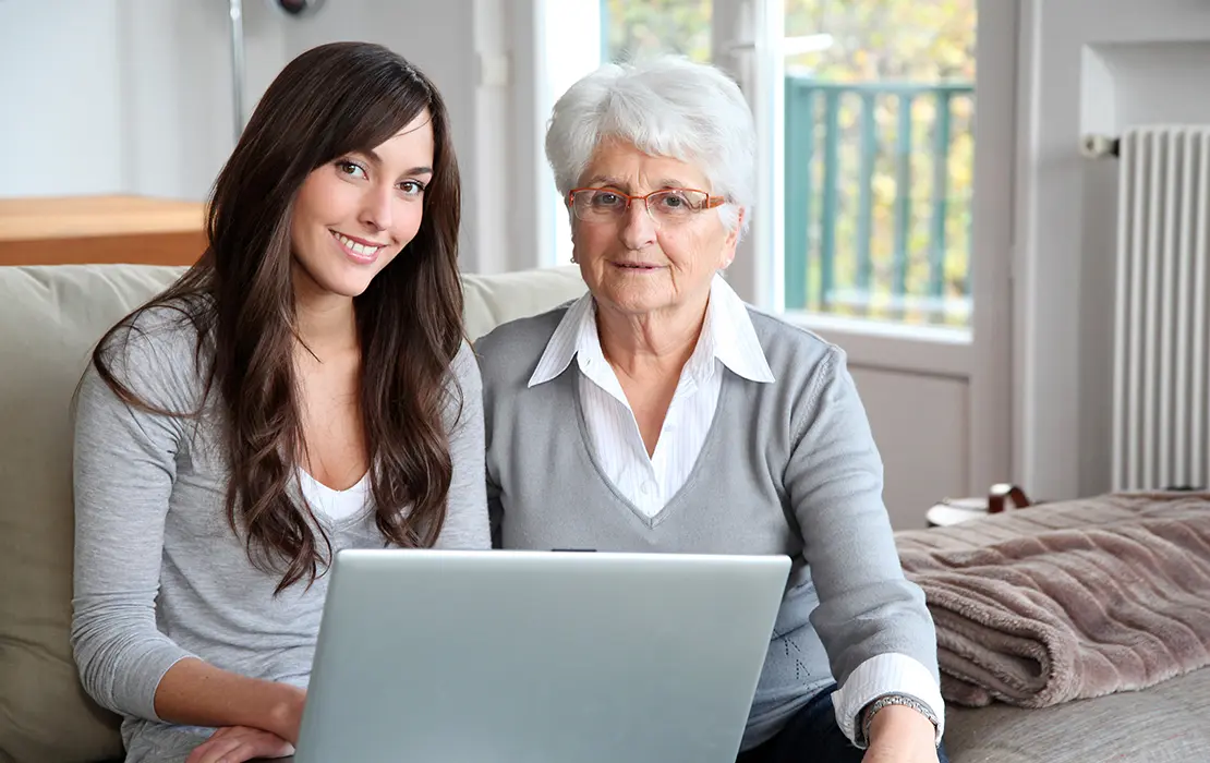 Pflegerin mit einer alten Dame auf einer Couch mit einem Laptop sitzend